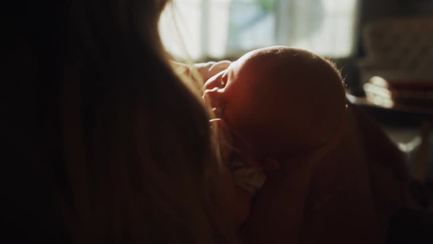 A loving mother gently breastfeeds her newborn baby, silhouetted by the warm light from a nearby window