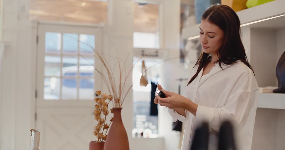 Stylish woman in a white blouse carefully sprays and smells a perfume sample on her wrist inside a bright boutique
