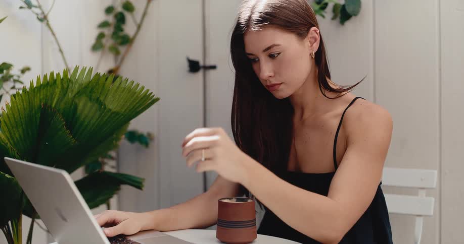 A thoughtful young woman works remotely on her laptop from a beautiful, plant-filled patio