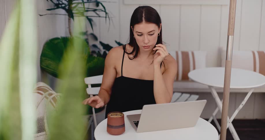 A stylish woman opens her laptop to work at a bright outdoor cafe before reaching into her bag for her wallet