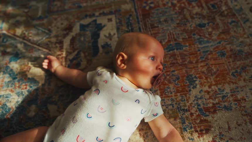 A cute newborn baby with a pacifier rests on a colorful patterned rug in the warm sunlight