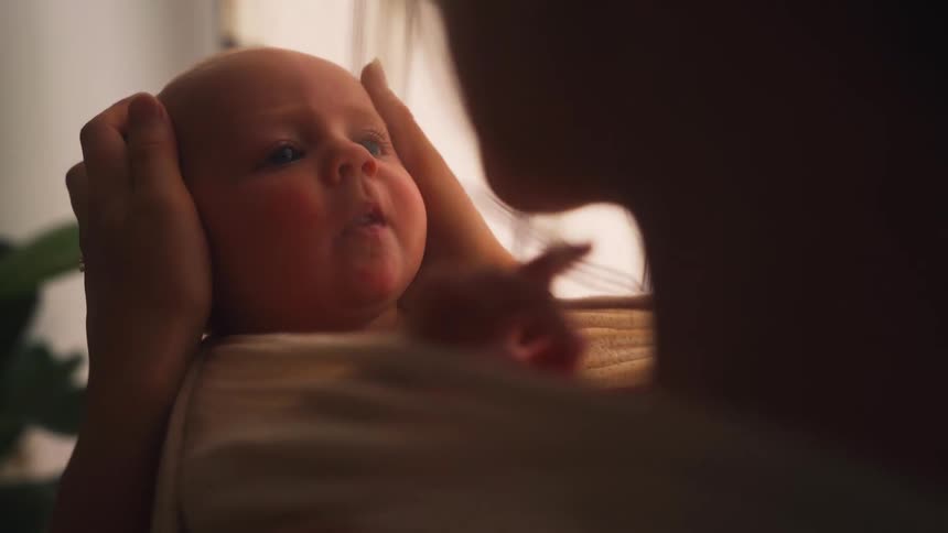 A mother cradles her newborn baby, who smiles and yawns in her arms in a softly lit room