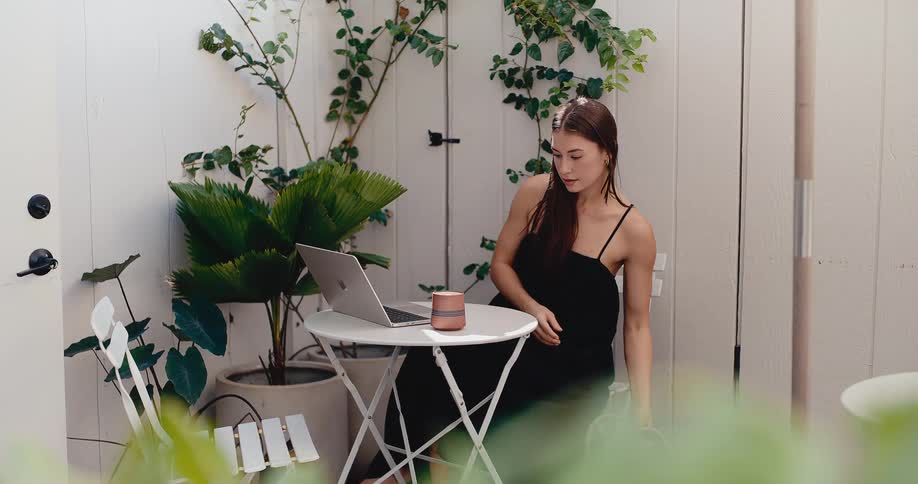 A young woman works on her laptop while sipping coffee on a serene, sunlit patio surrounded by lush green plants