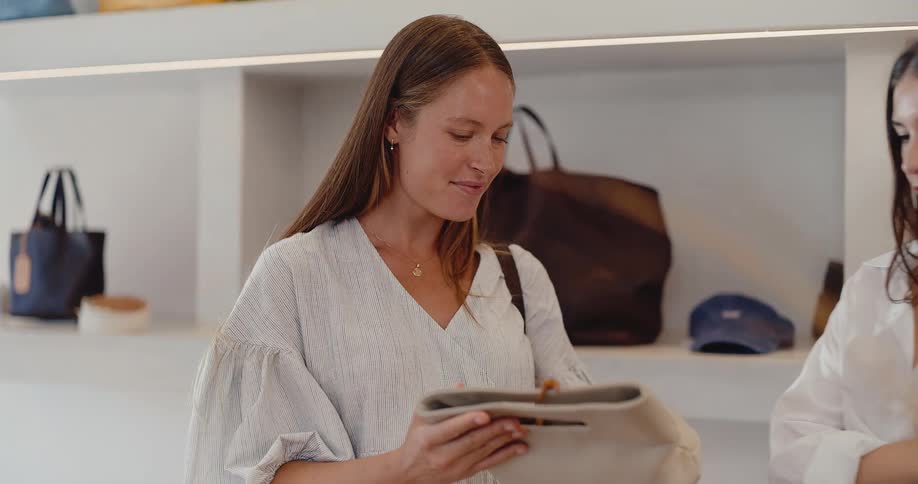 A smiling young woman carefully examines a stylish handbag with a friend or sales assistant in a modern, minimalist boutique