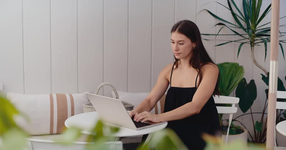 A young woman in a black dress works on her laptop while enjoying a coffee on a beautiful, bright patio surrounded by plants