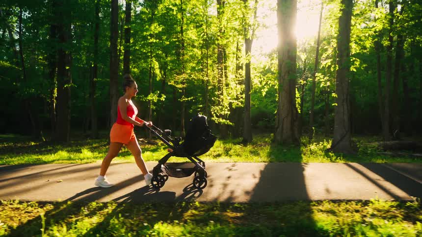 An athletic mother pushes a baby stroller along a sunlit path while exercising in a lush green park