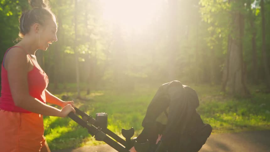 A smiling mother enjoys a beautiful golden hour walk while pushing her baby in a stroller through a lush park