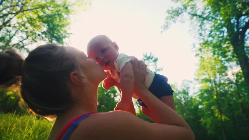 A mother joyfully lifts her newborn baby into the sunlit sky and kisses them in a beautiful green forest