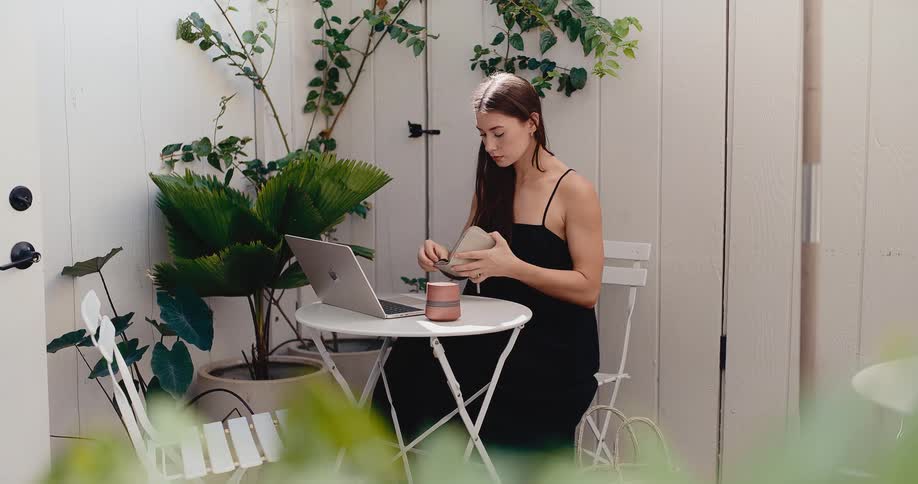 A stylish woman in a black dress uses her credit card to shop online on her laptop while sitting at a table on a bright, modern patio