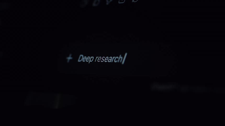 A user types the phrase 'Deep research about' into an artificial intelligence prompt on a computer screen in a dark room