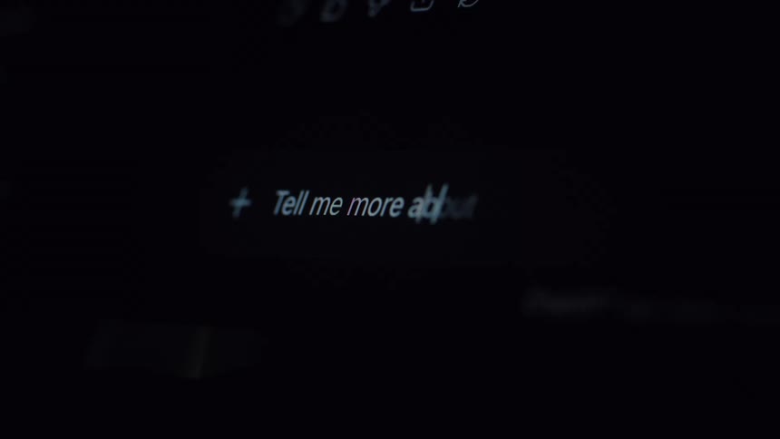 Close-up of a person typing a query 'Tell me more about...' into an AI chatbot on a dimly lit computer screen