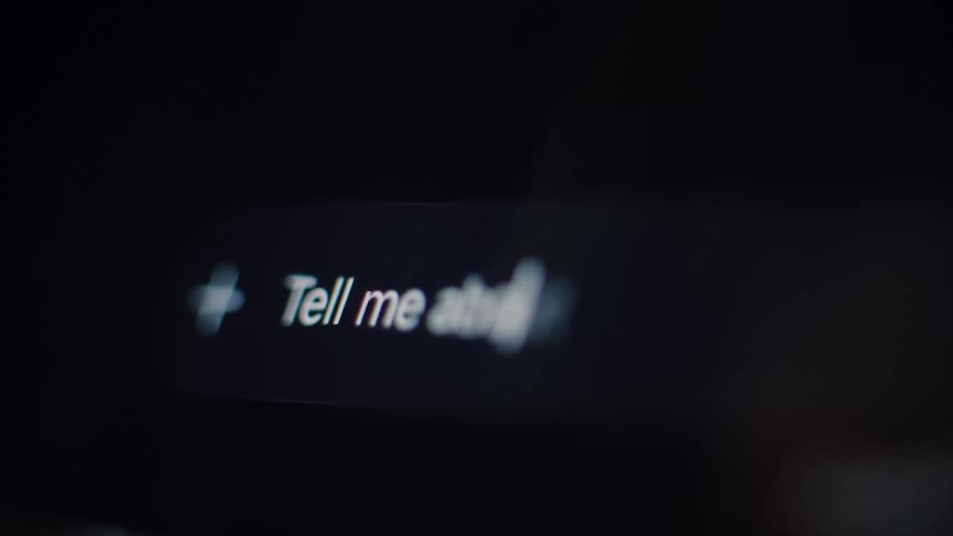 A close-up, out-of-focus shot of a user typing 'Tell me about...' into a search bar on a dark screen