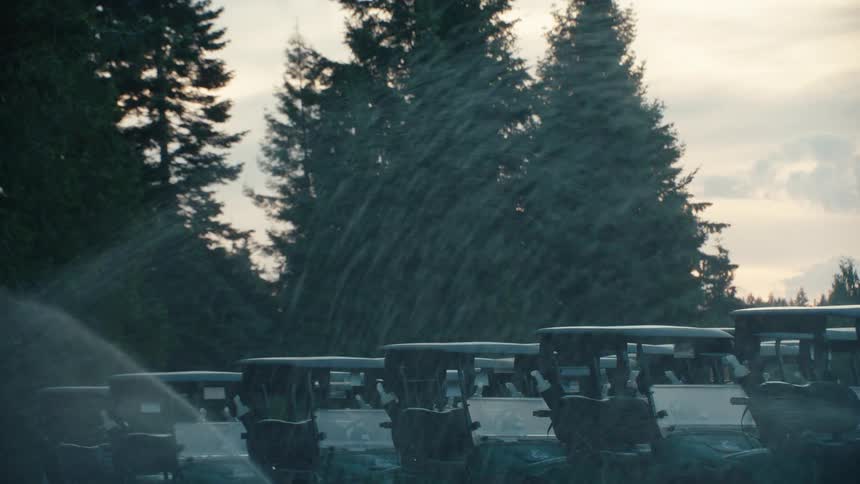 A sprinkler sprays water over a fleet of parked golf carts in slow motion at sunset