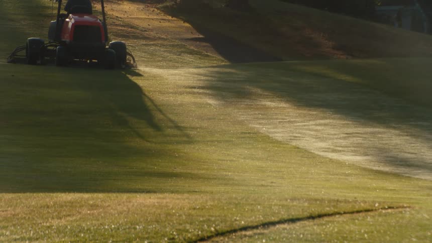 A groundskeeper operates a riding mower on a dewy golf course fairway in the beautiful morning light