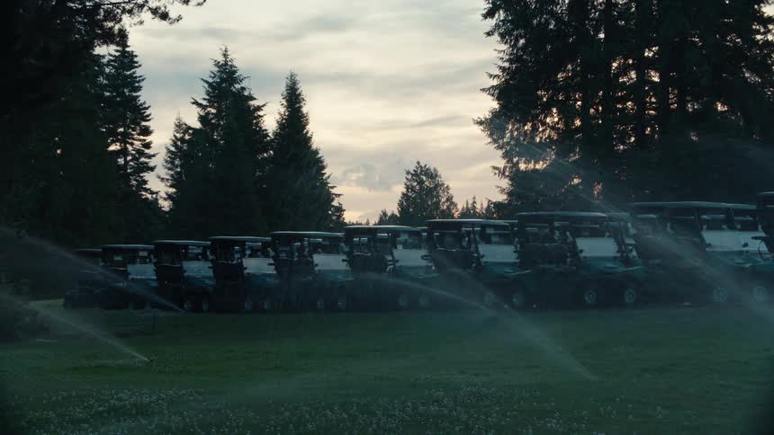 Sprinklers water a row of empty golf carts parked on a lush green course at dusk