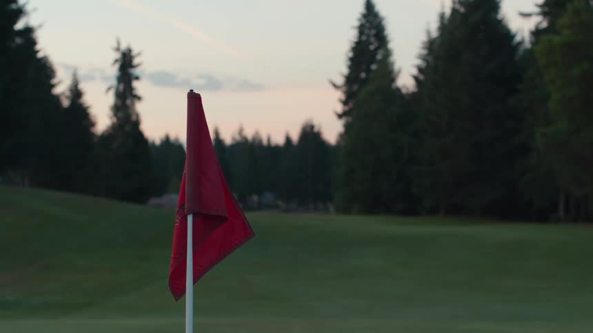 A close-up of a still red golf flag on a tranquil course with a backdrop of evergreen trees at dusk