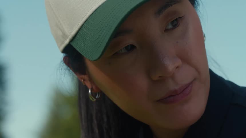 A focused Asian woman wearing a baseball cap and hoop earrings concentrates intently on a sunny day