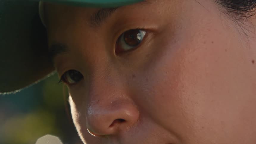 Sunlit extreme close-up of a young Asian person in a baseball cap looking up thoughtfully