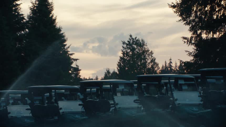A sprinkler waters a row of parked golf carts at dusk on a quiet golf course