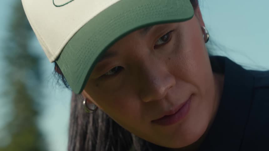 An extreme close-up of a focused Asian woman in a baseball cap concentrating on her golf game on a sunny day