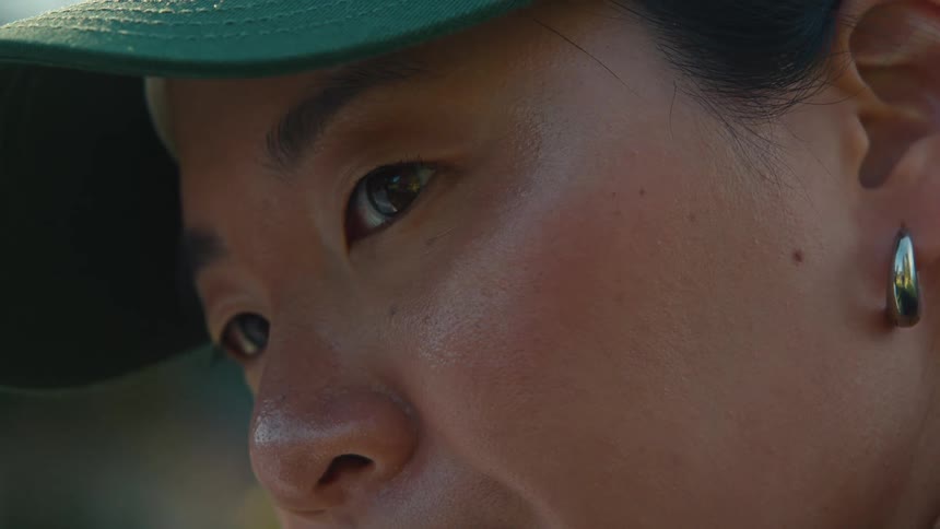 An intimate close-up shot of an Asian person's face, focusing on their eye, earring, and cap as they concentrate on something off-screen