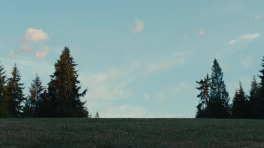 A low-angle shot shows a groundskeeper driving a large riding lawn mower over a grassy knoll against a scenic backdrop of trees and sky