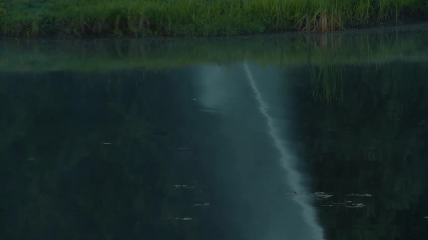 Ethereal fog drifts across the tranquil, dark waters of a lake, reflecting the lush green shoreline at dusk