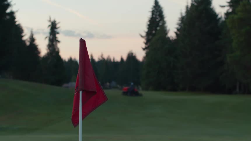 A red golf flag stands still on a dewy green as a groundskeeper mows the course early in the morning