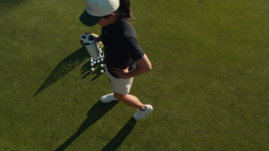 A high-angle, slow-motion view shows a female golfer walking away from a basket of practice balls and a putter on a lush green