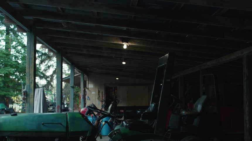 A static shot of old farm equipment parked in a dimly lit rustic open-air garage