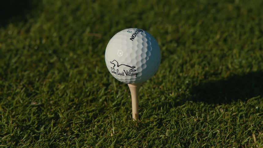 A tattooed hand carefully places a golf tee and ball onto the lush green grass of a golf course in a close-up shot