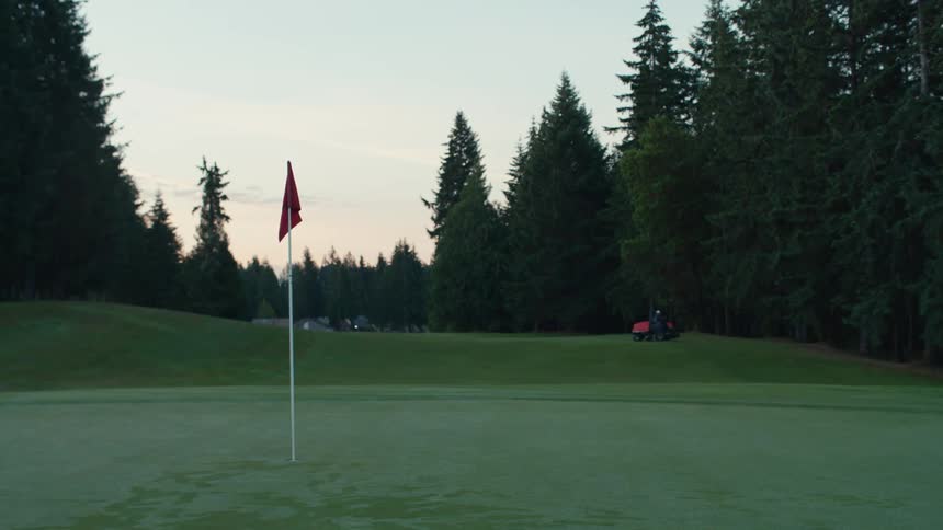 Early morning light illuminates a pristine golf course green as a groundskeeper on a riding mower maintains the fairway in the background