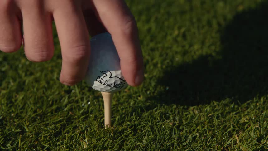 A slow-motion close-up shot of a hand carefully placing a golf ball on a wooden tee on a lush green course