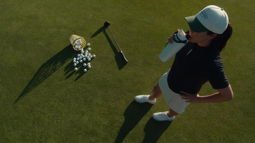 Top-down view of a woman golfer taking a break from practice to drink from her water bottle on a sunny green
