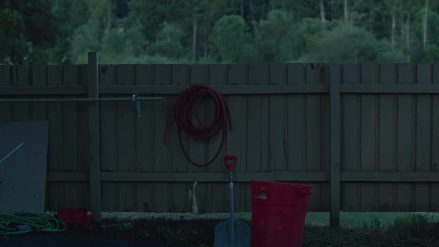 A still, dark shot of a backyard fence with a coiled red hose, shovel, and trash can at dusk