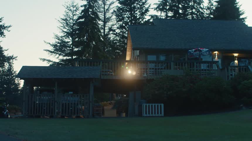 A golf cart pulls up to a cozy cabin-style house with a brightly lit deck at dusk