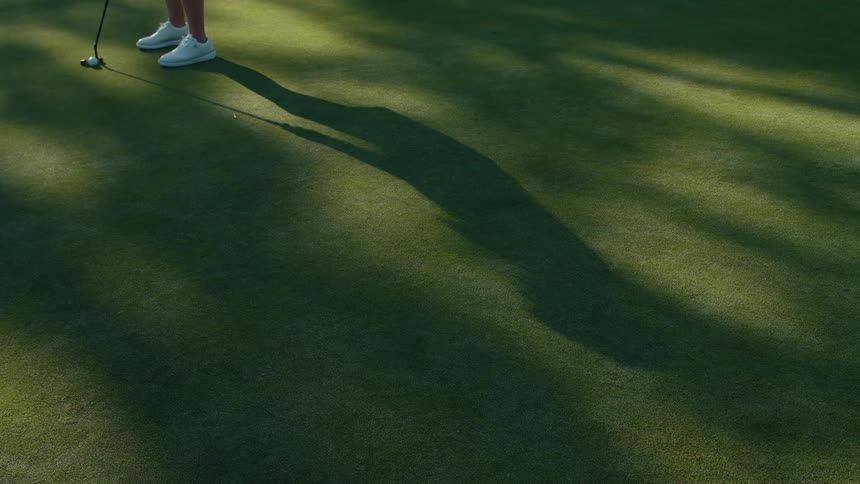 A golfer's long shadow is cast across the green as they putt the ball, narrowly missing the hole in slow motion