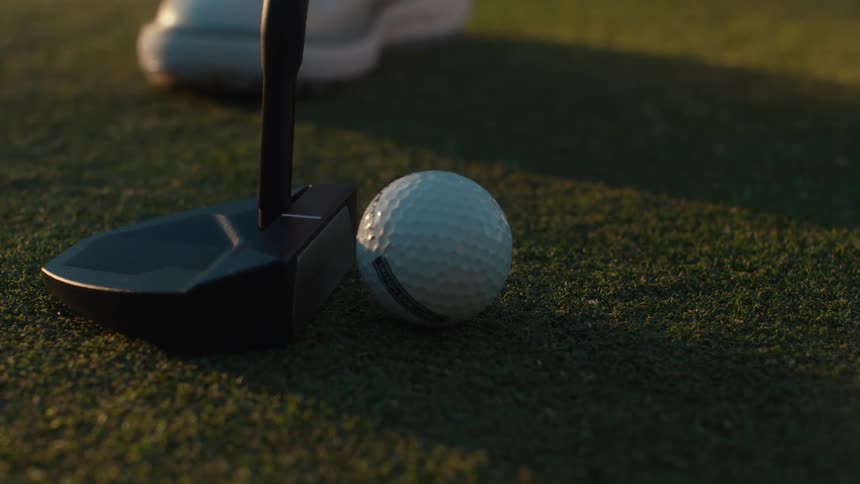 A golfer's putter strikes a golf ball in slow motion on a manicured green during sunset
