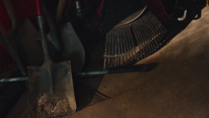 A collection of well-used shovels and rakes lie on a concrete floor in a dimly lit garage