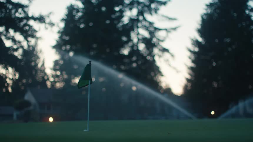 A golf ball rolls onto a putting green near the flagstick as sprinklers water the course at dusk