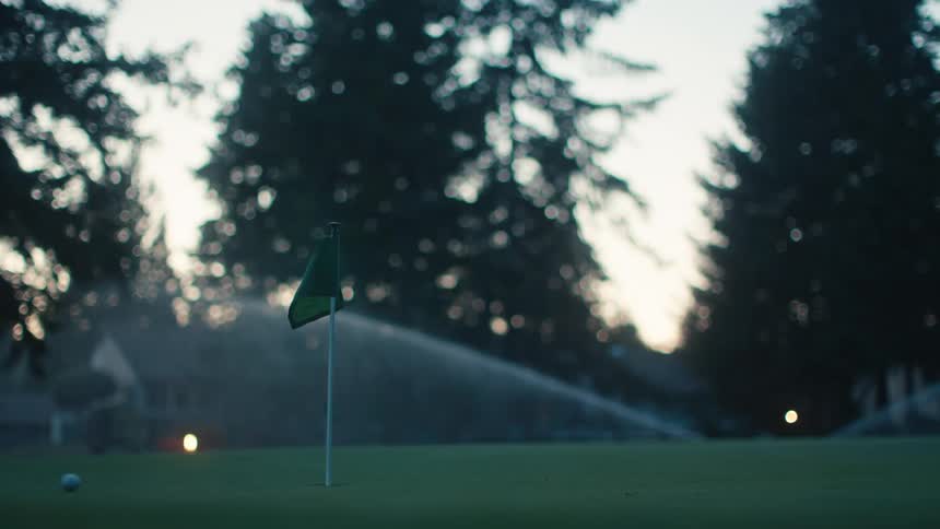 A golf ball slowly rolls to a stop inches from the flag on a manicured green as sprinklers water the course at dusk