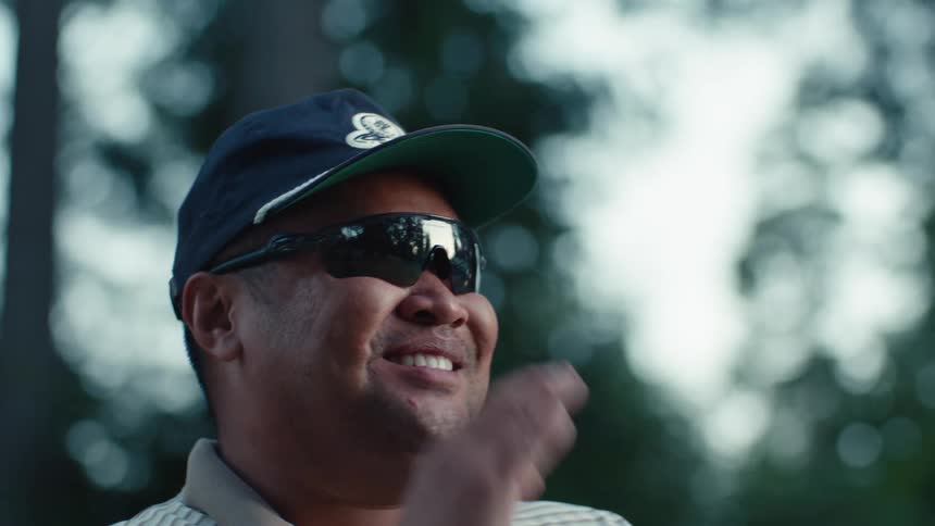 A man wearing a baseball cap and sunglasses smiles and talks outside in a park