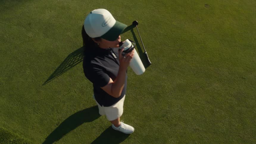 High-angle slow-motion view of a woman golfer pausing for a drink of water during a practice session on a sunny green