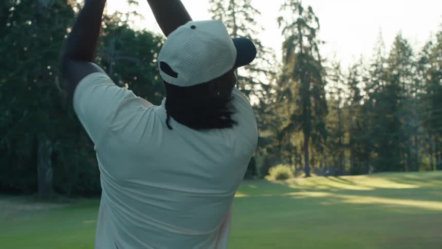 A man with dreadlocks takes a powerful swing with a golf club in slow motion on a green course