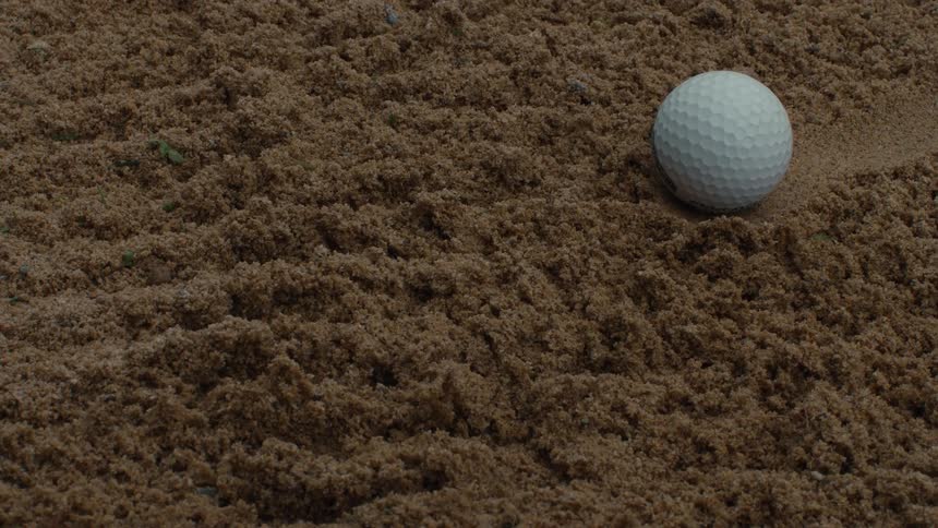 A close-up static shot of a single white golf ball resting in a brown sand trap