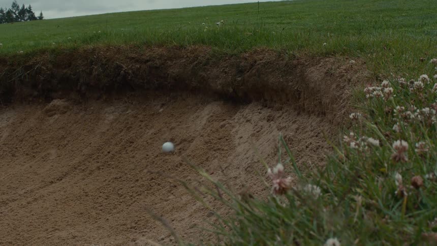A golf ball is hit into a sand bunker, landing on the steep face and rolling down into the sand in slow motion