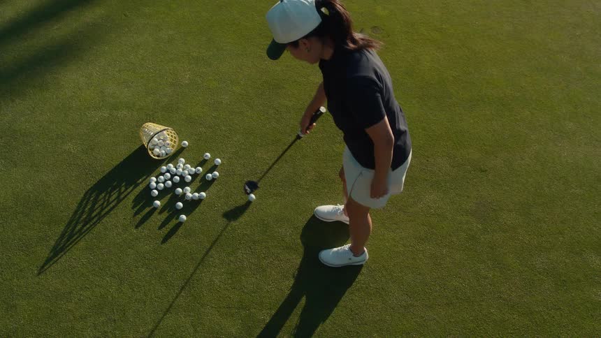 A high-angle slow-motion shot shows a female golfer practicing her putting on a pristine green with a basket of balls