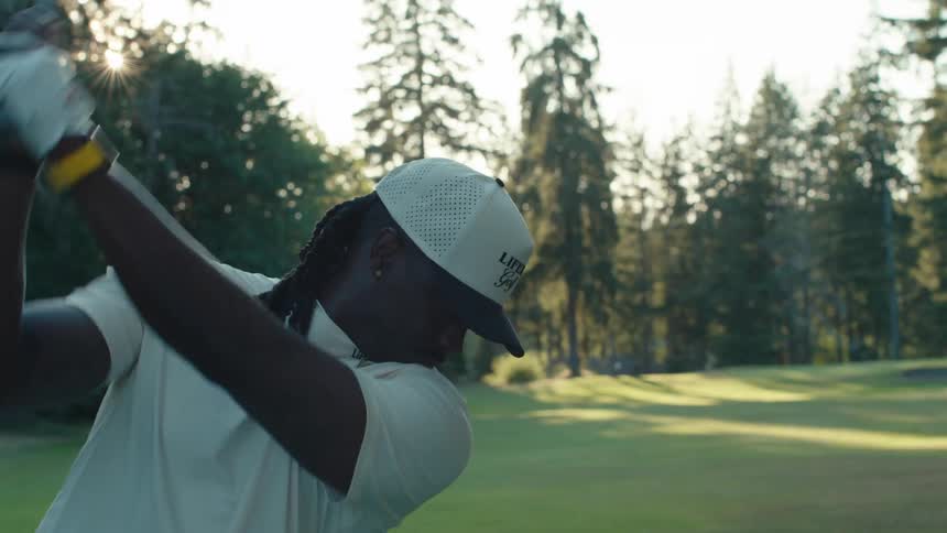 A golfer with dreadlocks swings his club in slow motion on a lush green course backlit by the sun