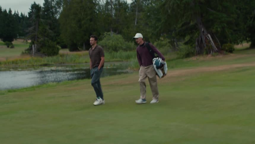 Two men walk and talk together on a lush green golf course next to a pond