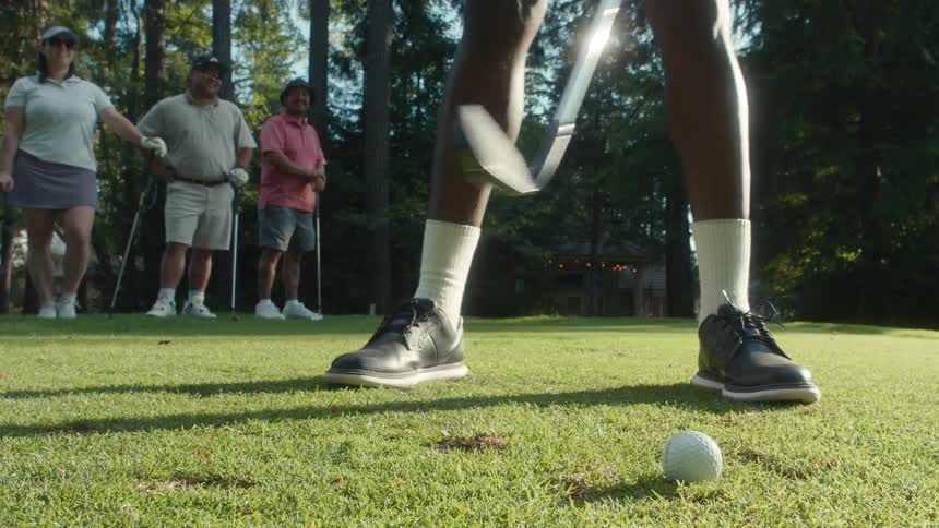 A slow-motion, low-angle view shows a golfer hitting a ball off the grass as friends watch on a sunny course